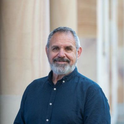 A man with greying hair and beard wearing a navy shirt stands with sandstone columns behind him.